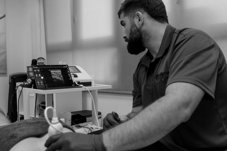 Ander Blanco explora con la sonda del ecógrafo la espalda de un paciente, con el equipo en el carro; en blanco y negro