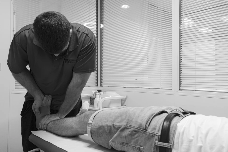 Ander Blanco moviliza la pierna de un paciente tumbado en la camilla, en una sala con ventanas y estores; en blanco y negro
