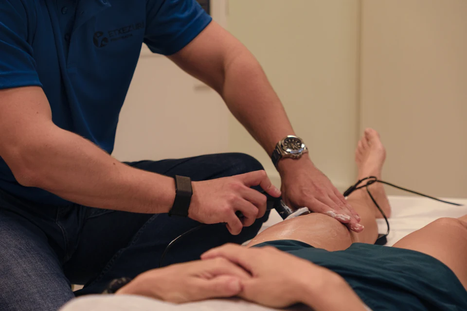 Manos del fisioterapeuta deslizando un cabezal con gel sobre el muslo de una paciente, que apoya las manos entrelazadas