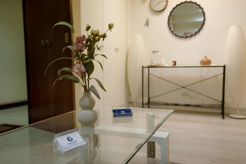 Sala de espera de Etxezuri: mesa de cristal con el tarjetero de la clínica, jarrón con flores rosas, consola negra, lámparas de papel y espejo redondo