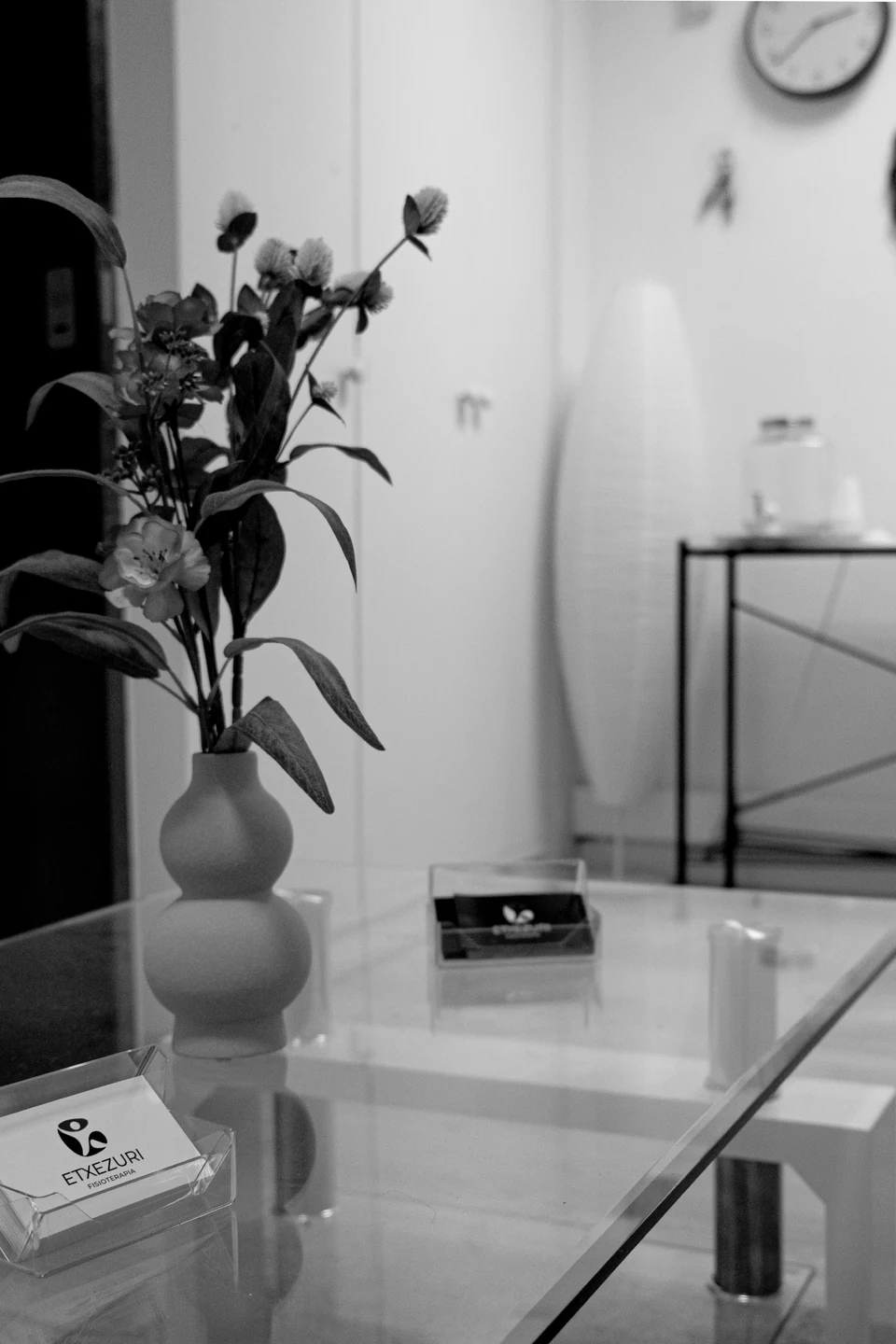 Mesa de cristal de la sala de espera de Etxezuri con un jarrón de flores y el tarjetero con las tarjetas de la clínica; en blanco y negro
